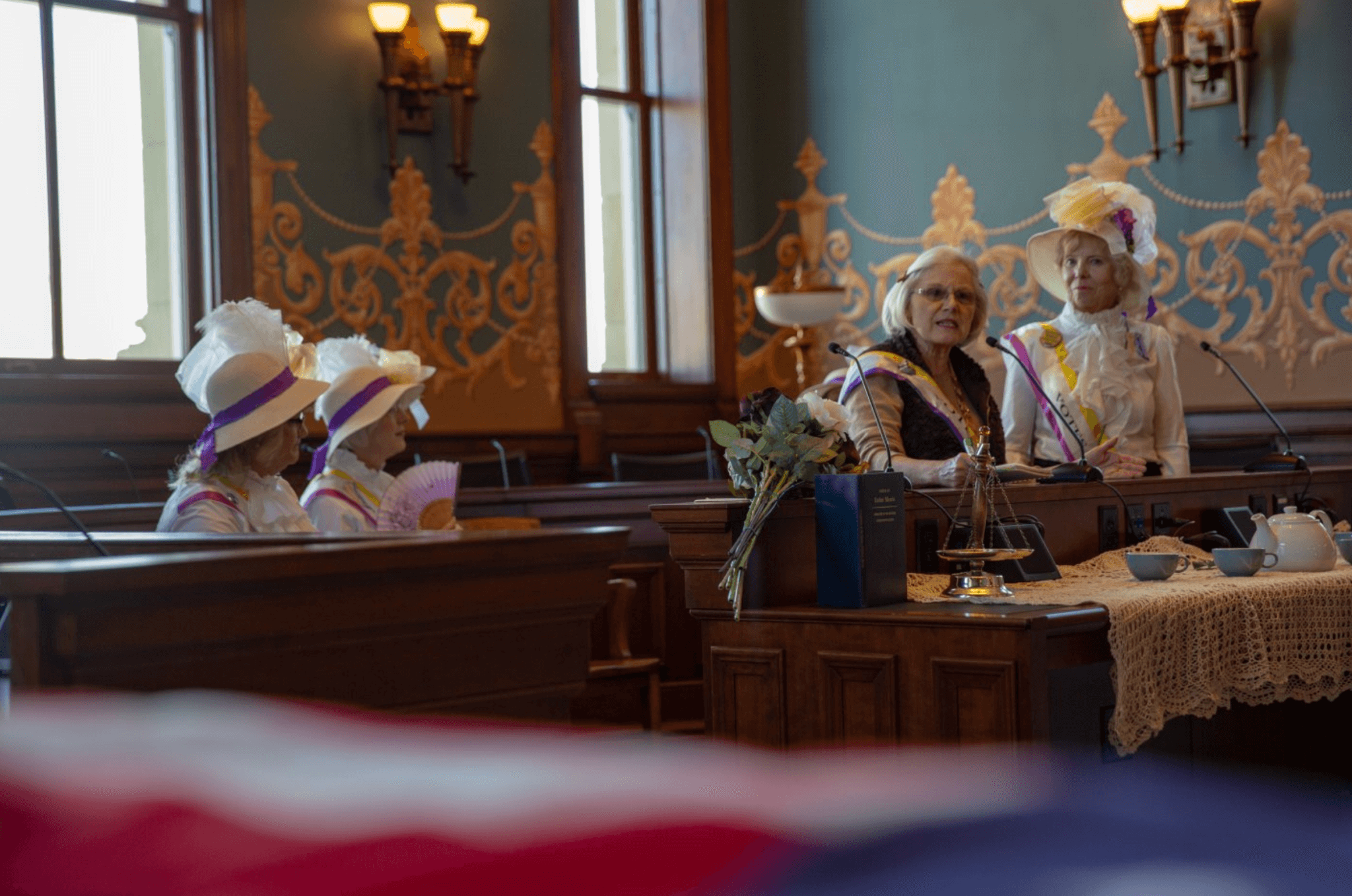 Women wearing suffrage sashes that read 'Vote' gather in a historic building, seated before teacups.